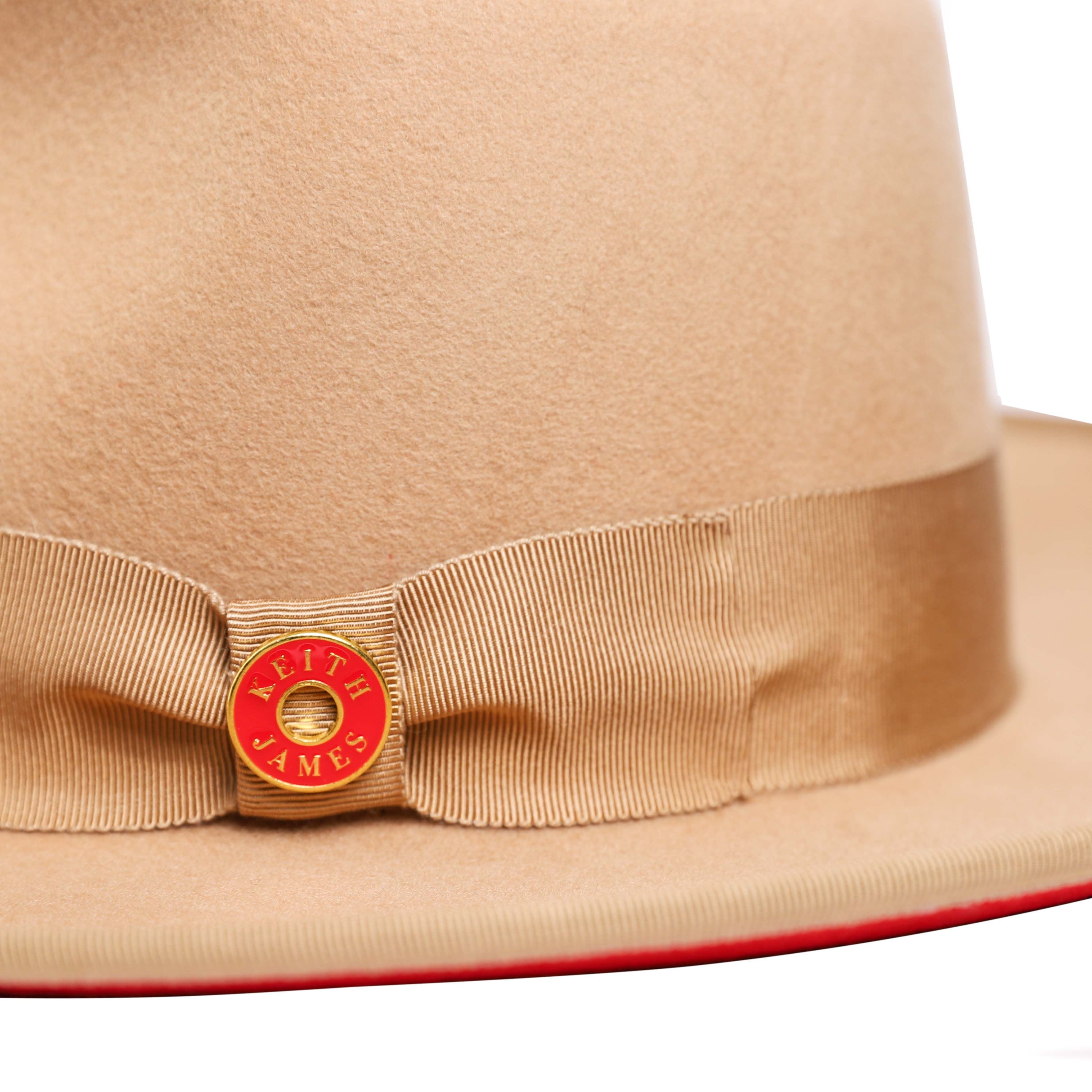 A golden honey colored fedora hat with a wide brim and a signature logo pin on the band.