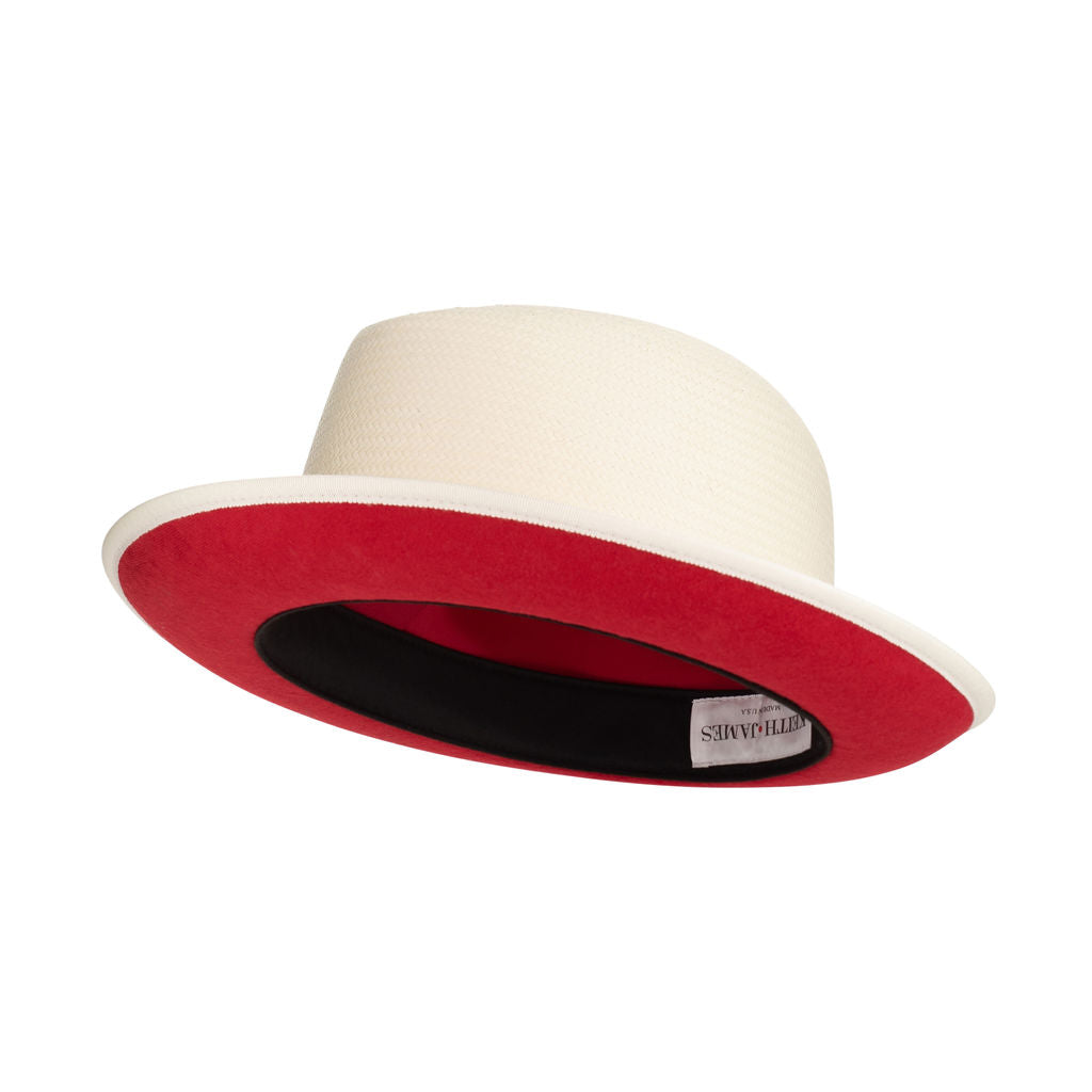 A beige classic fedora hat with a red bottom brim and a signature logo pin.