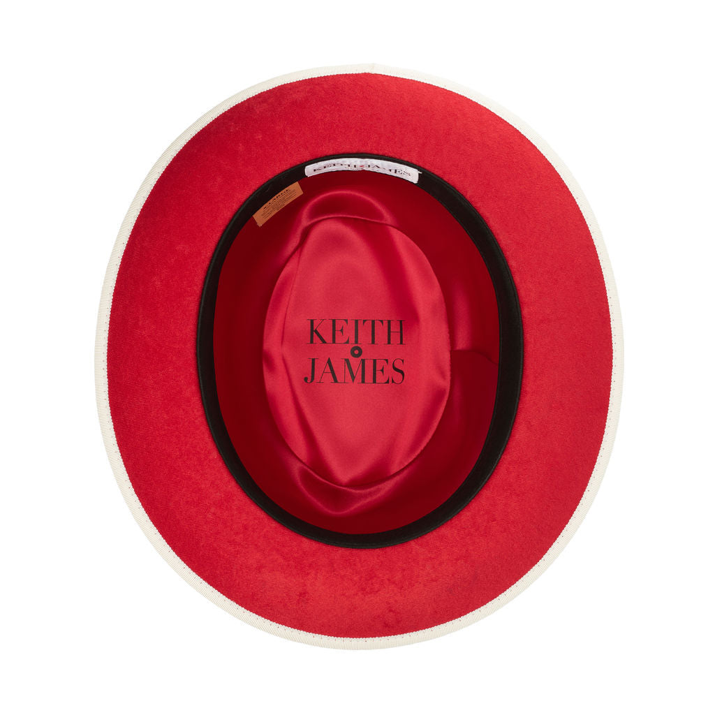 A beige classic fedora hat with a red bottom brim and a signature logo pin.