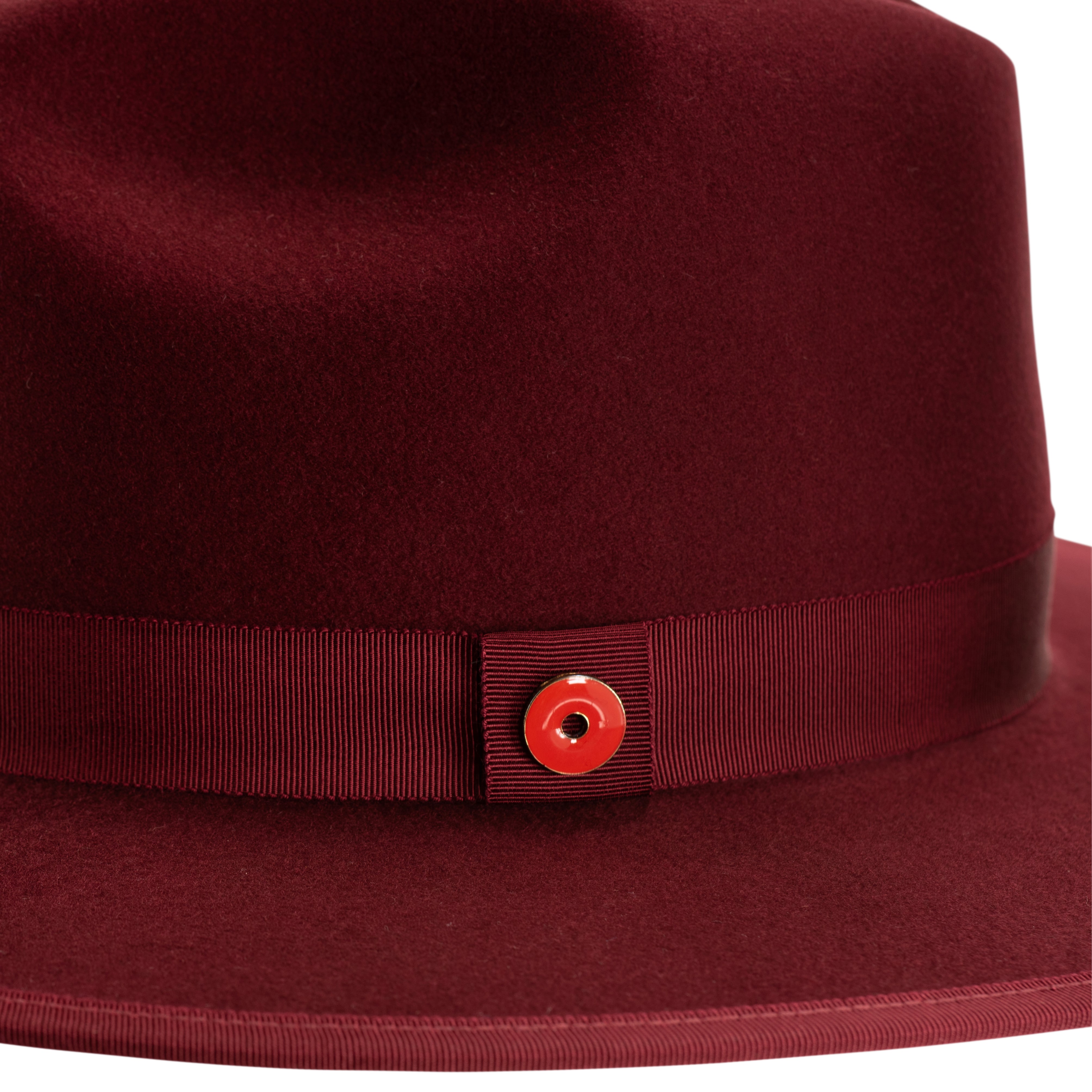 A maroon colored fedora hat with a flat brim and a classic ribbon trim, featuring a signature logo pin on the side.