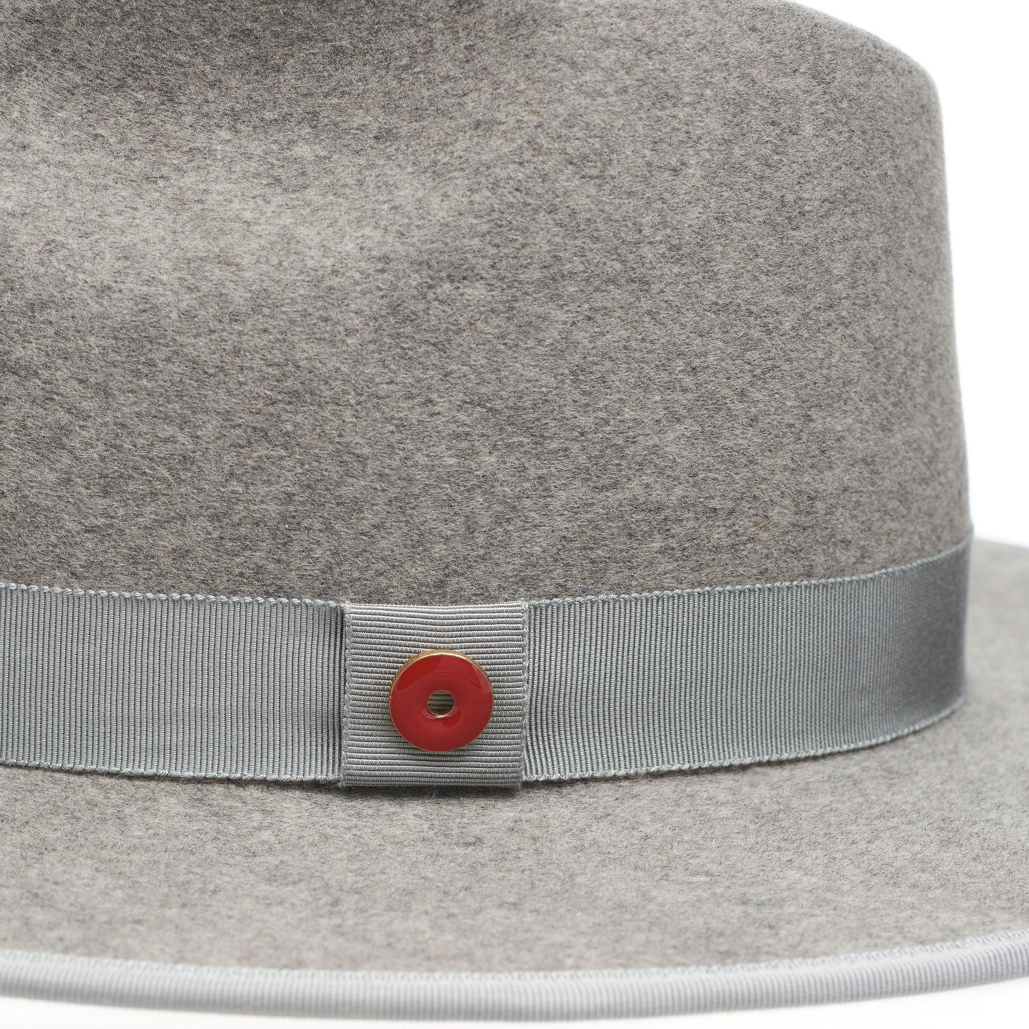 A grey fedora hat with a flat brim and a classic ribbon trim, featuring a signature logo pin and a red bottom brim.
