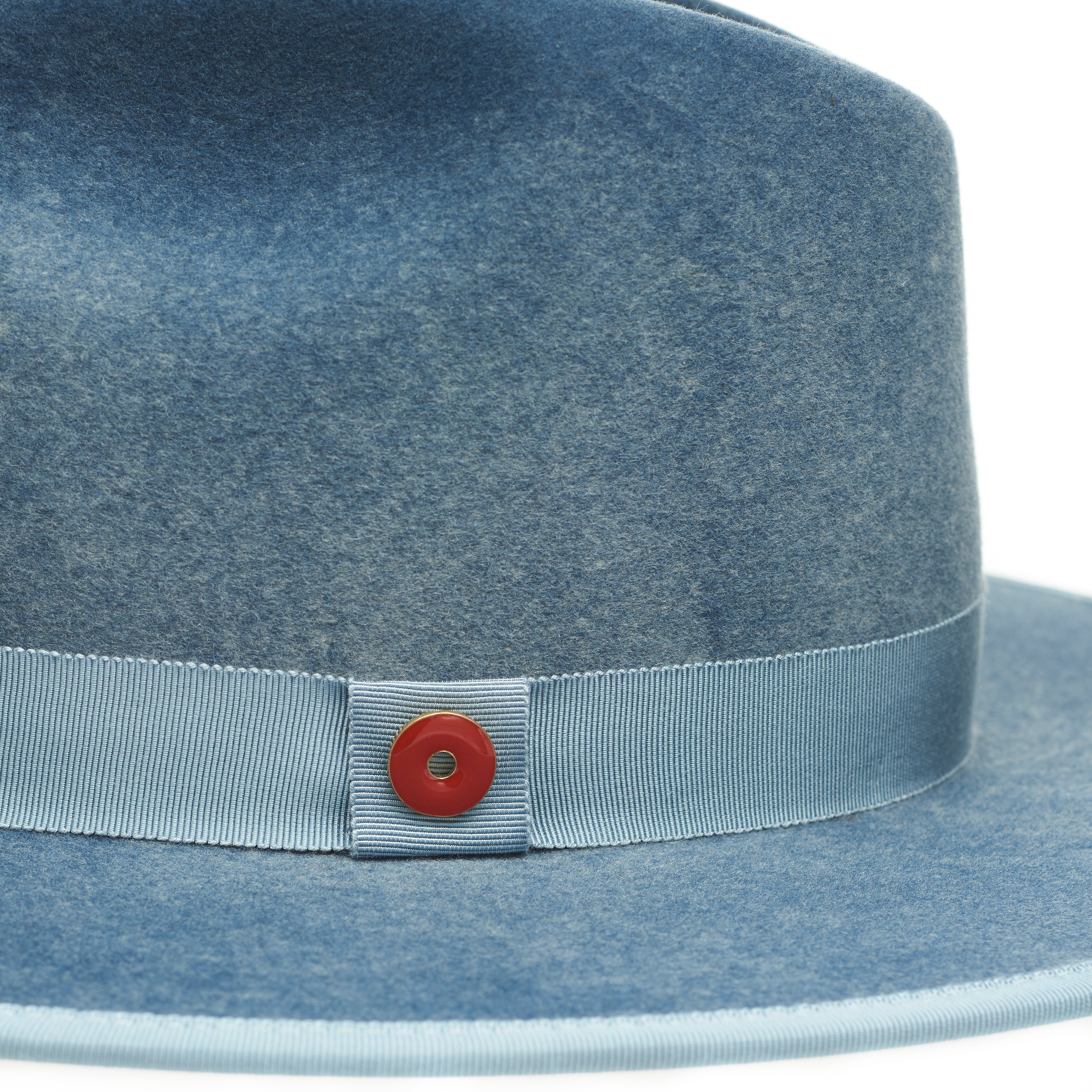 A blue fedora hat with a flat brim and a classic ribbon trim, featuring a signature logo pin and a red bottom brim.