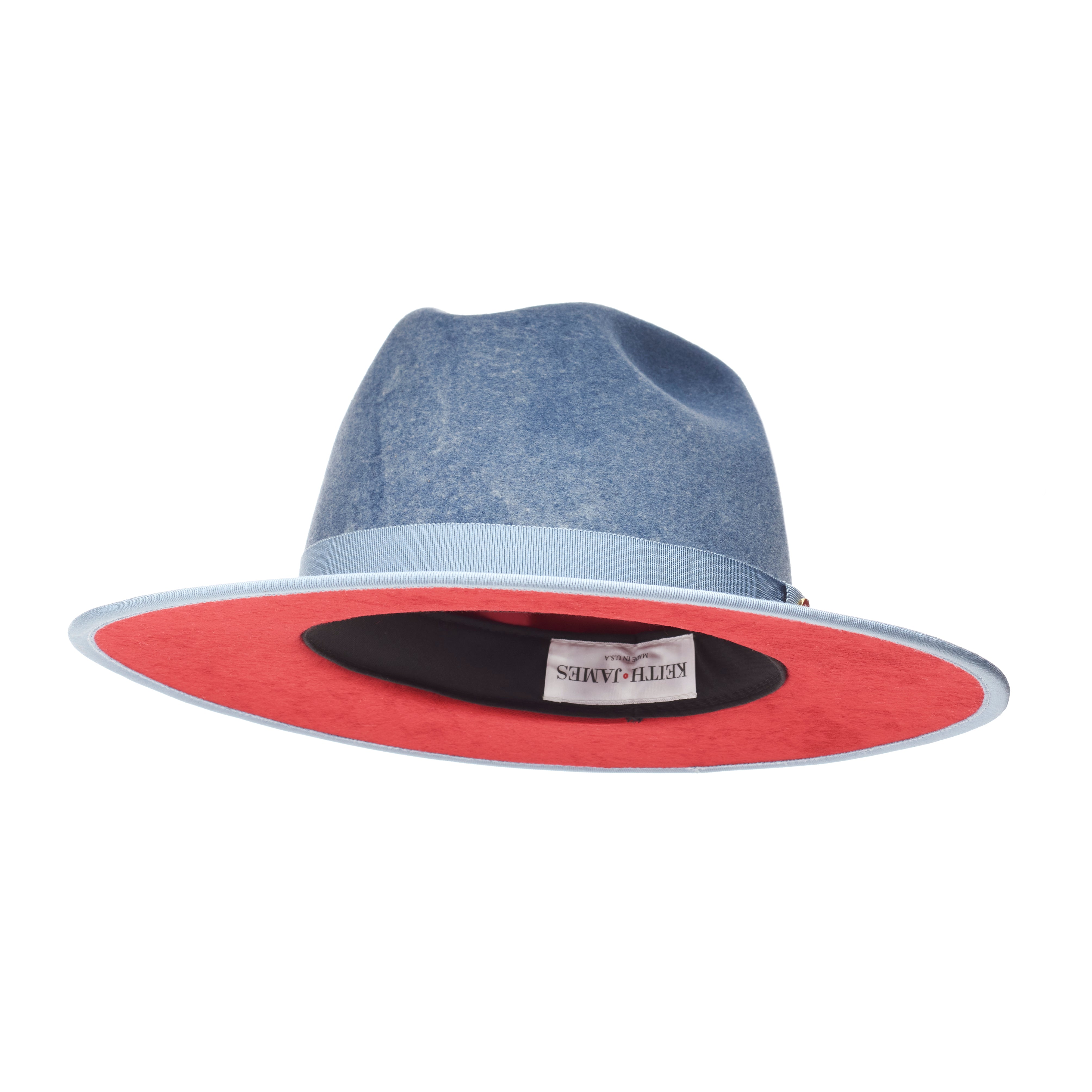 A blue fedora hat with a flat brim and a classic ribbon trim, featuring a signature logo pin and a red bottom brim.