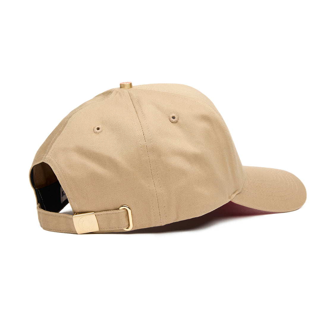 A beige 5 panel baseball cap with an adjustable band and a logo embroidered on the front, featuring a red bottom brim.