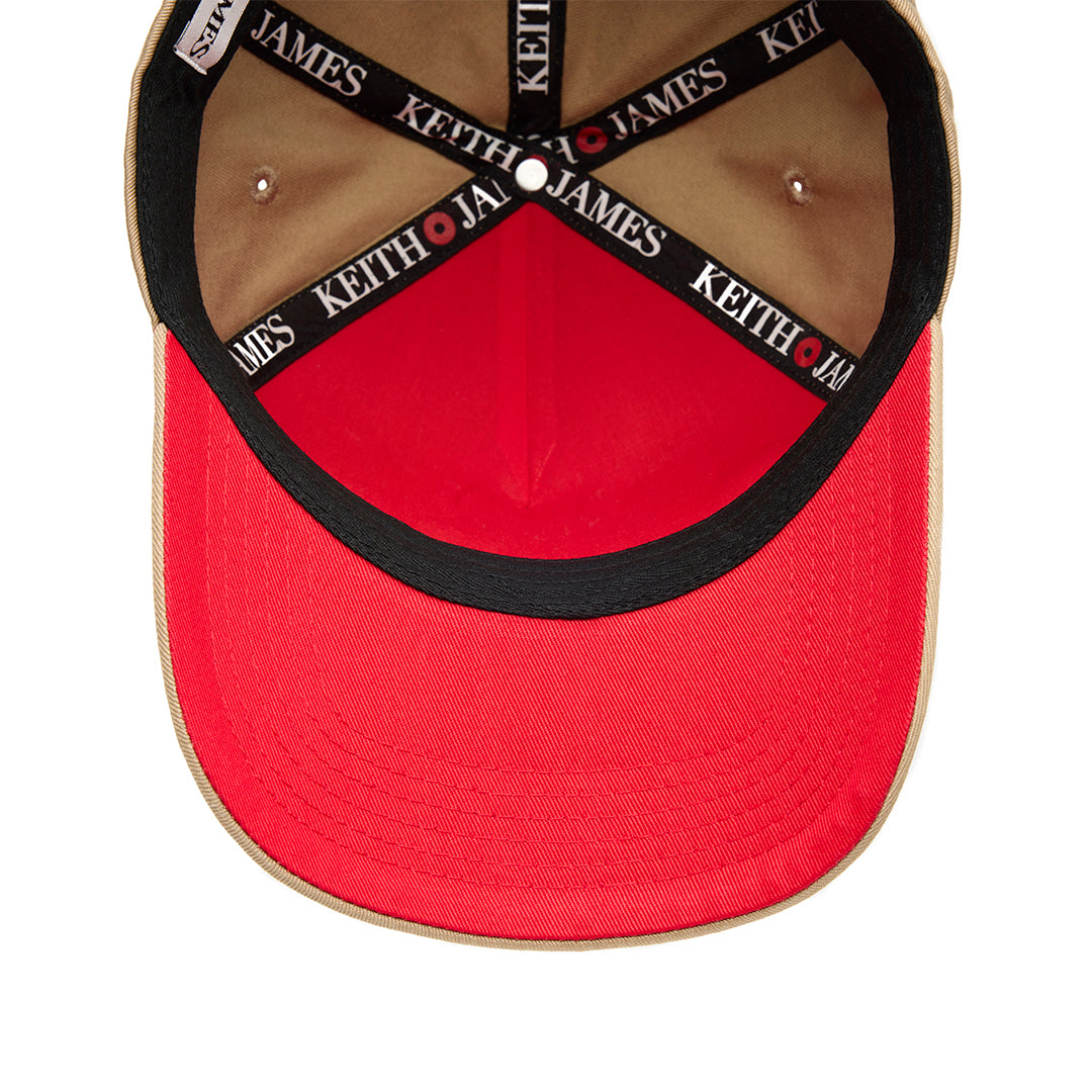 A beige 5 panel baseball cap with an adjustable band and a logo embroidered on the front, featuring a red bottom brim.