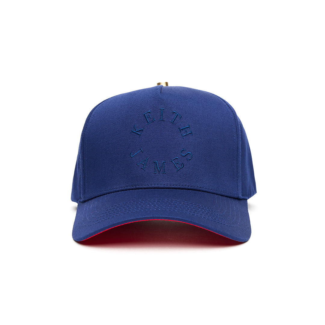 A navy blue 5 panel baseball cap with an adjustable band and a logo embroidered on the front with a red bottom brim.