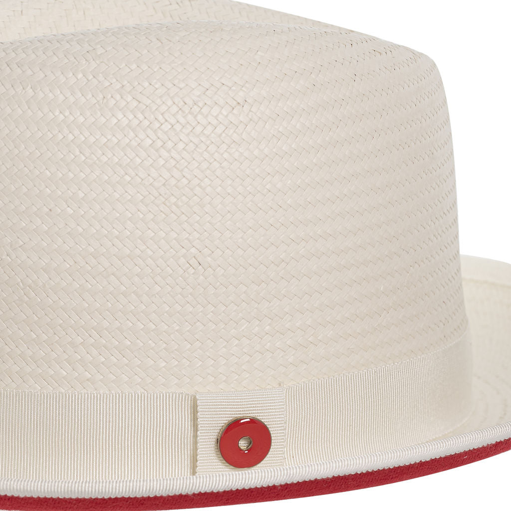 A beige classic fedora hat with a red bottom brim and a signature logo pin.