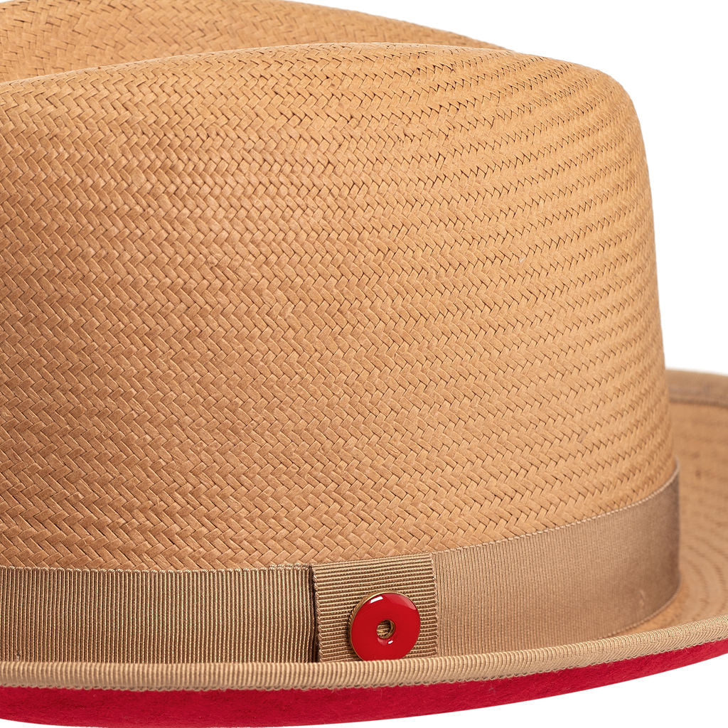 A golden honey straw fedora hat with a red bottom brim and a signature logo pin.