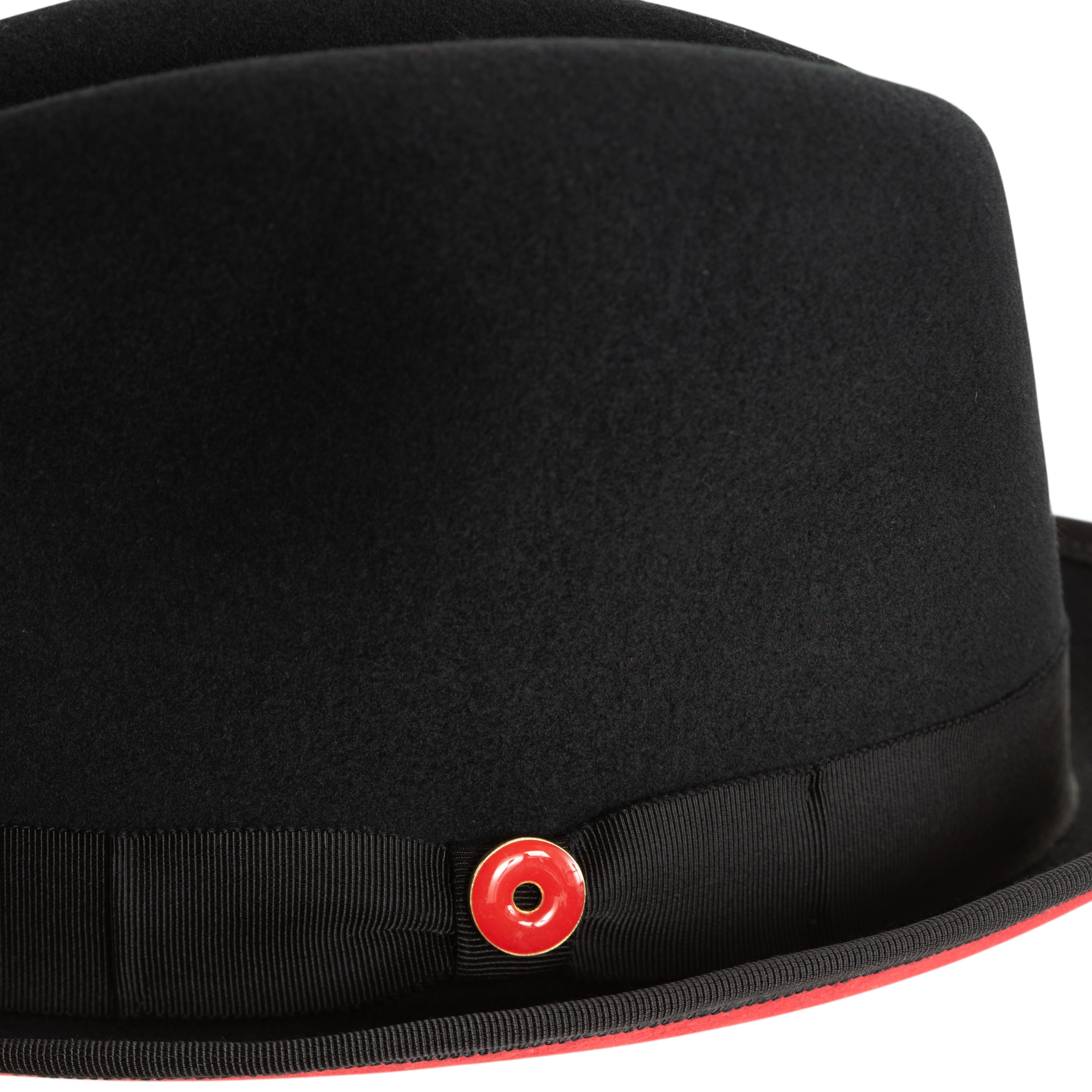 A black pinched crown fedora hat with a red bottom brim and a signature logo pin.