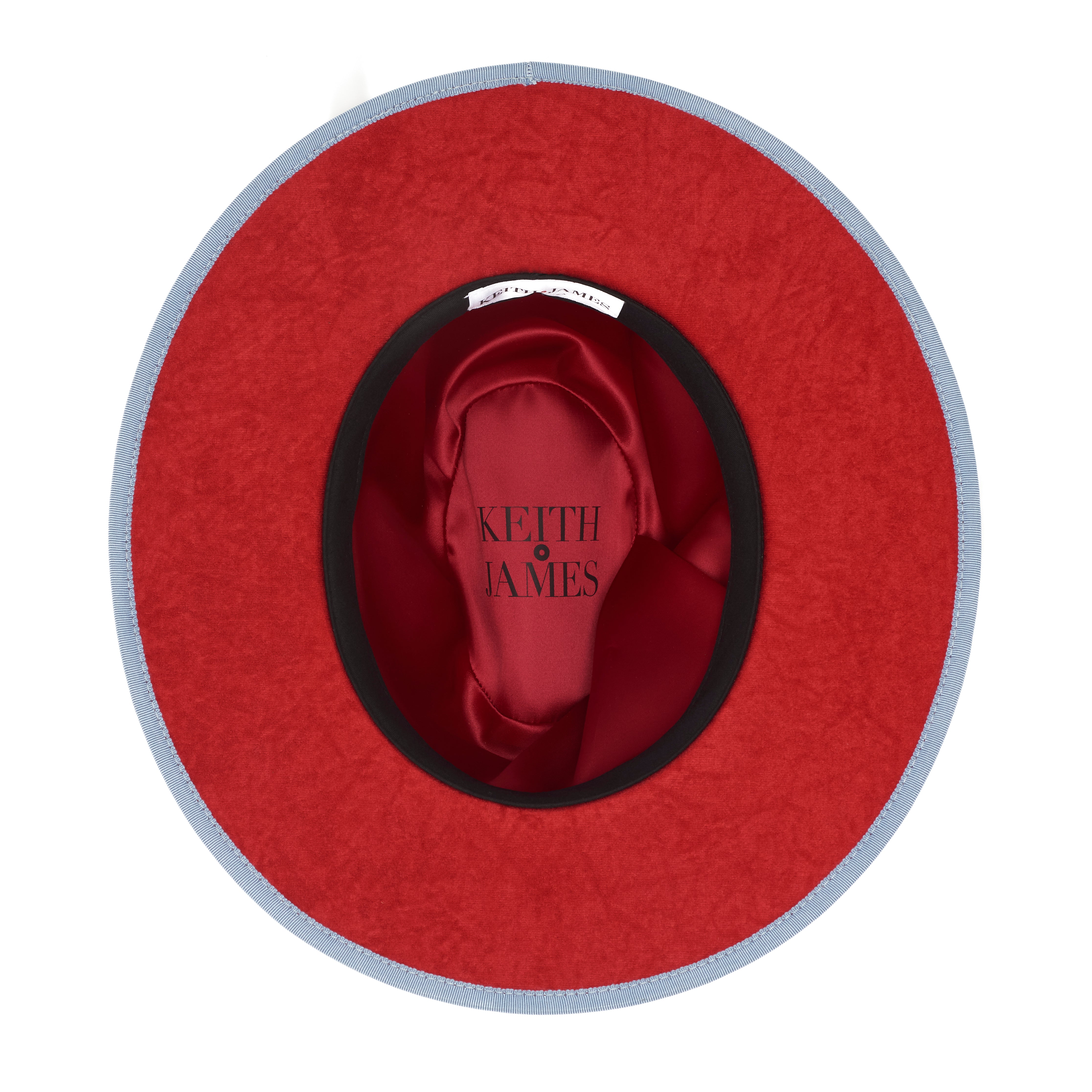 A blue pinched crown fedora hat with a flat brim and a classic ribbon trim, featuring a signature logo pin and a red bottom brim.