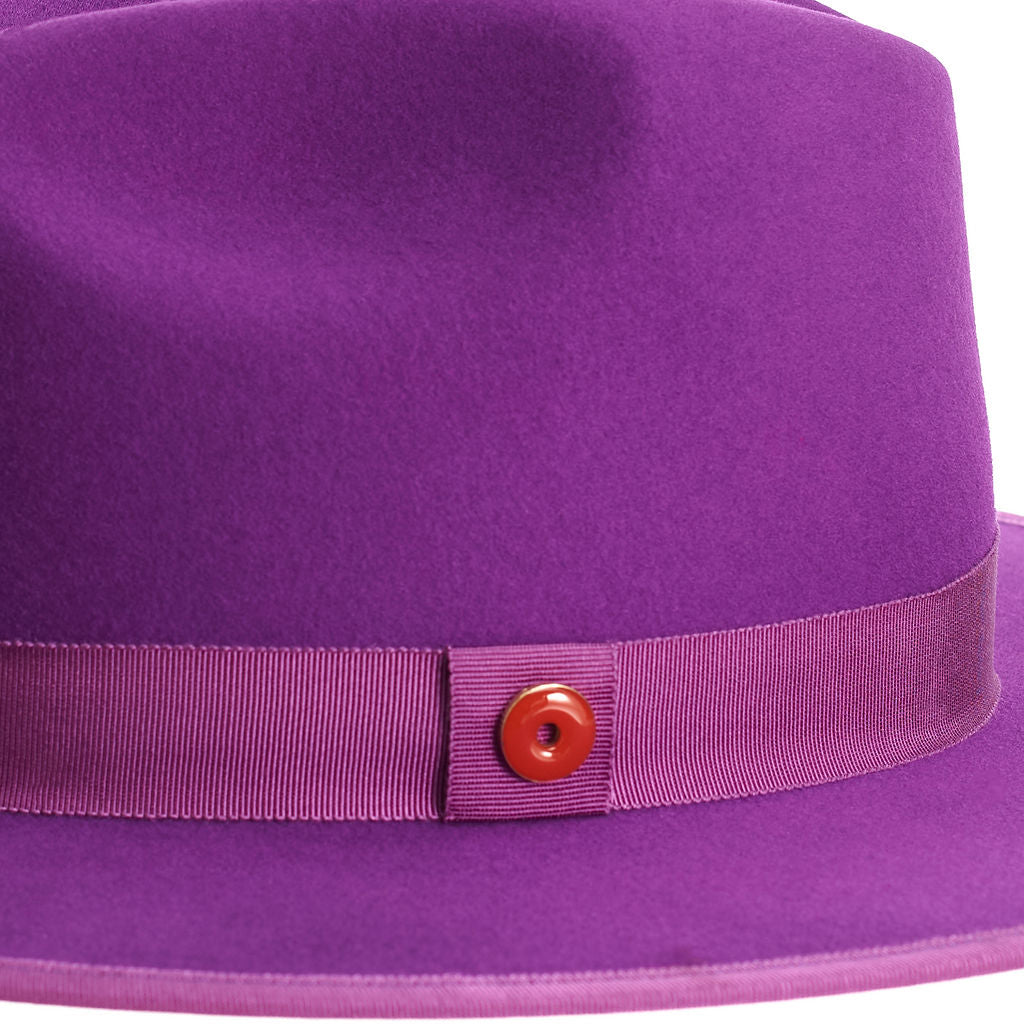 A violet purple fedora hat with a flat brim and a classic ribbon trim, featuring a signature logo pin and a red bottom brim.
